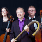 Vasemmalta oikealle naissellisti, mies, joka pitää kädessään isoa sauvaa sekä miessellisti seisomassa tumman taustan edessä.