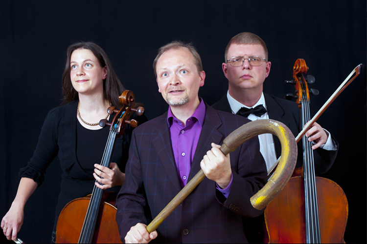 Vasemmalta oikealle naissellisti, mies, joka pitää kädessään isoa sauvaa sekä miessellisti seisomassa tumman taustan edessä.