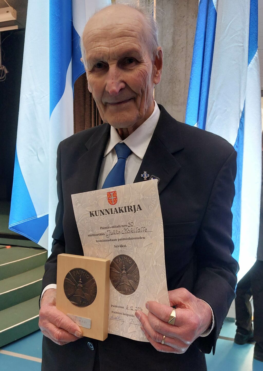 Tummaan pukuun pukeutunut mies pitää kädessään Paimio-mitalia ja sen kunniakirjaa. Taustalla lippujalkoihin asetettuja Suomen lippuja.