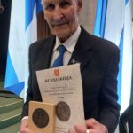 Tummaan pukuun pukeutunut mies pitää kädessään Paimio-mitalia ja sen kunniakirjaa. Taustalla lippujalkoihin asetettuja Suomen lippuja.