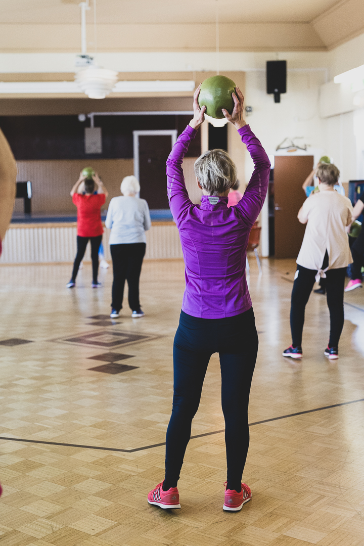 Nainen violetissa paidassa pitää käsissään vihreää palloa jumppatunnilla