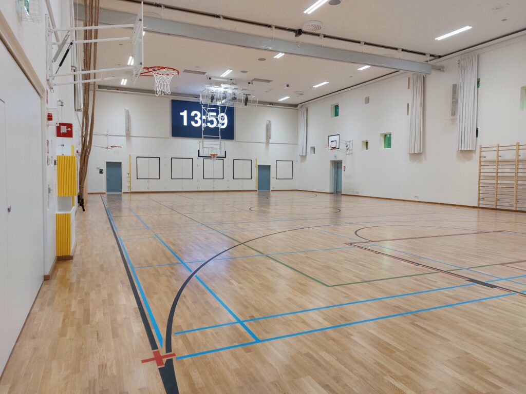 Puulattiainen liikuntasali. Takaseinällä iso tv-taulu, jossa kellonaika 13.59.