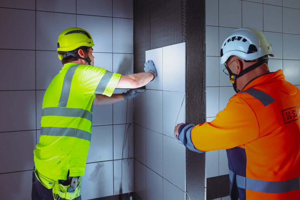Kaksi kypäräpäistä miestä laatoittamassa seinää.