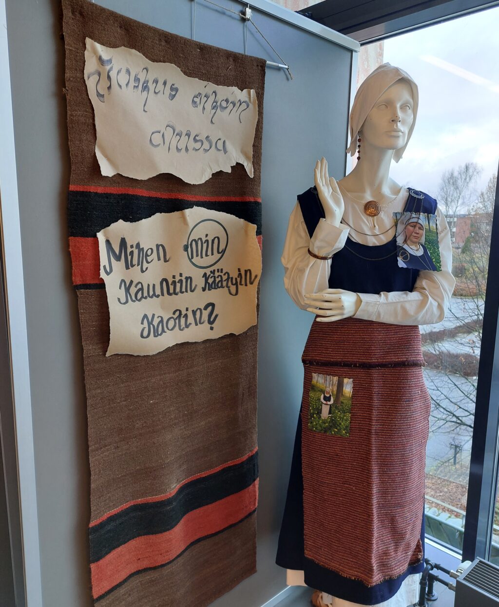 Näyttelysenäkkeeseen ripustettu tekstiili sekä mallinuken päällä naisen asusteita.