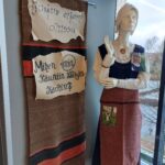 Näyttelysenäkkeeseen ripustettu tekstiili sekä mallinuken päällä naisen asusteita.