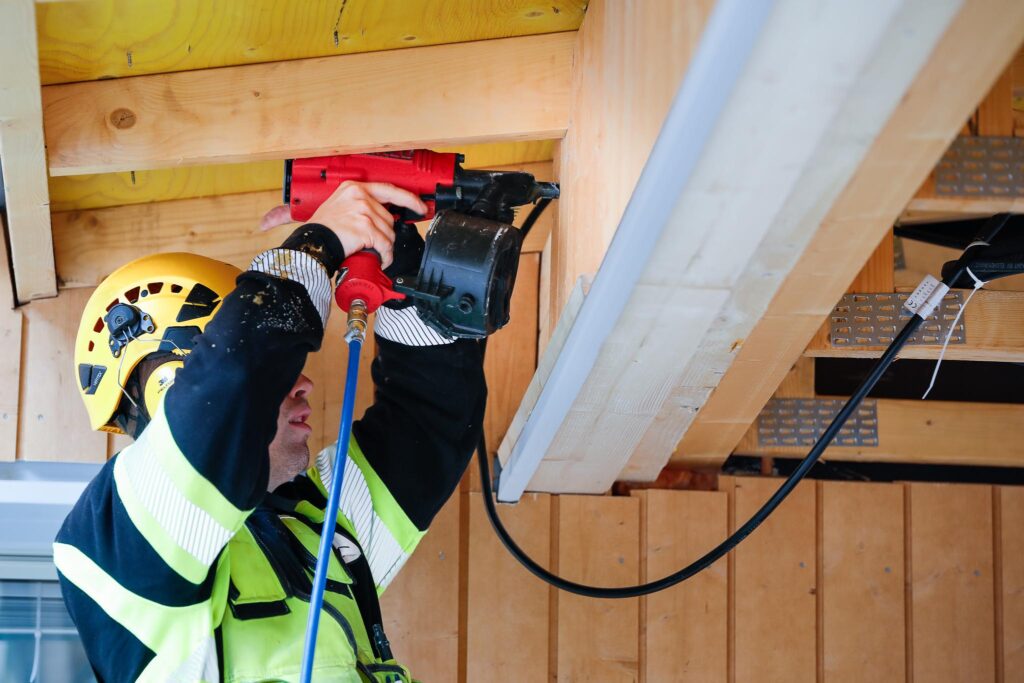 Mies keltaisessa kypärässä ja työasussa kiinnittaa jotakin rakennuksen vaakapalkkiin.