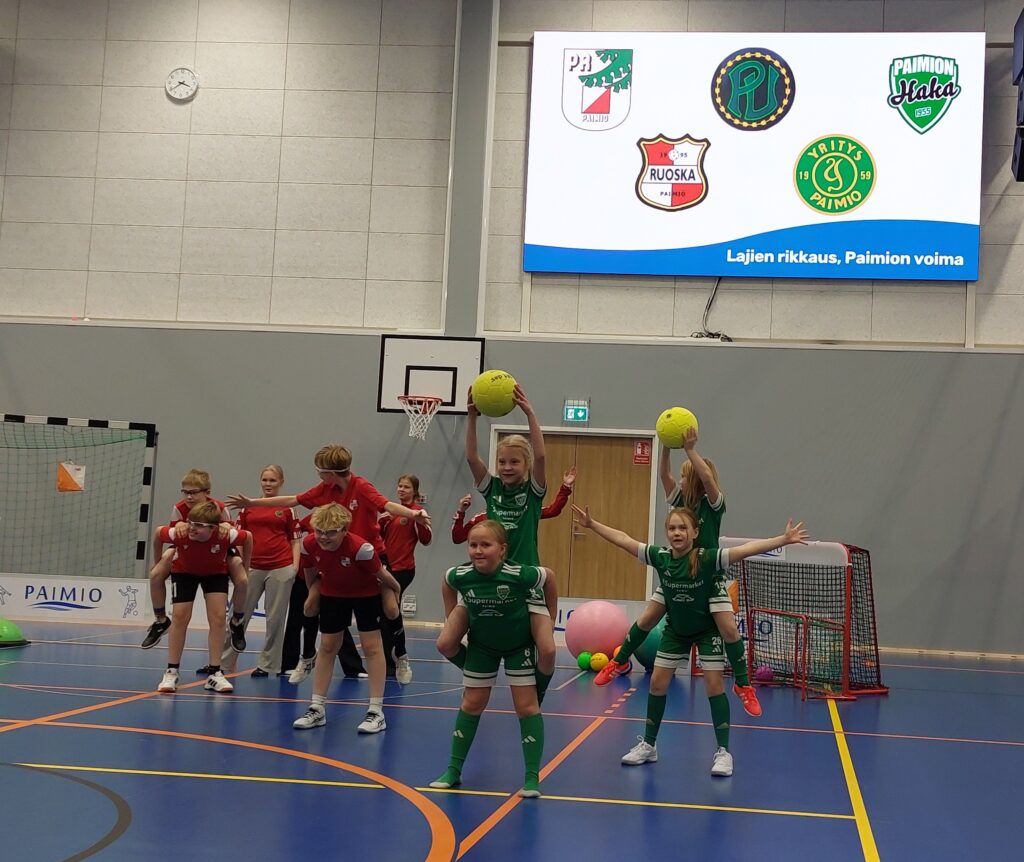 Urheiluasuisia, eri seurojen lapsia ja nuoria liikkumassa, sinipohjaisella sisäliikuntahallin lattialla. Taustalla seinällä iso tv-näyttö, jossa viiden paimiolaisseuran logot.