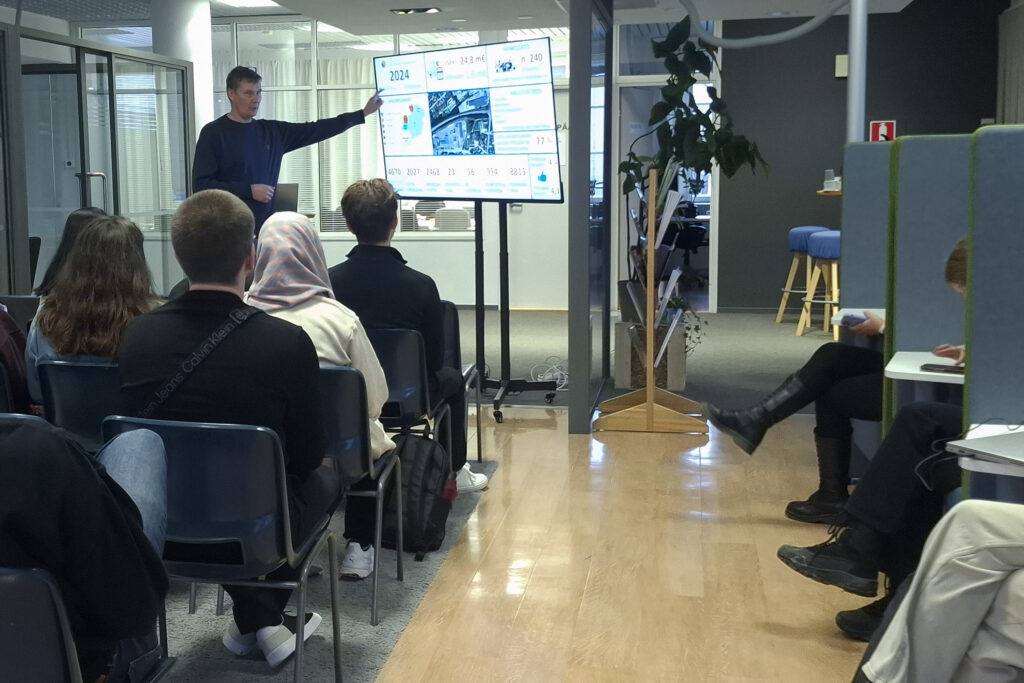 Ihmisiä seuraamassa mieshenkilön esitystä. Esittelijän vieressä tv-näyttö, johon esittelijä osoittaa kädellään.