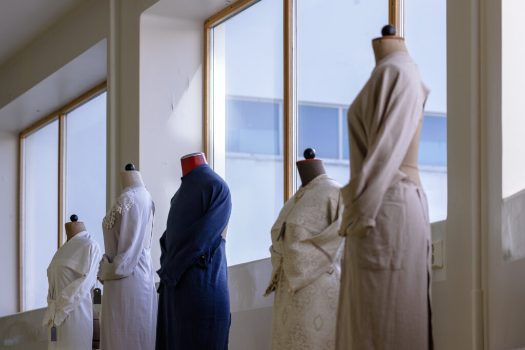 Viisi mallinukkea isojen ikkunoisen edessä. Kunkin nuken päällä erilainen vaate.