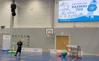 Isossa liikuntahallissa nainen puhuu mikrofoniin. Salin lattialla on liikuntavälineitä ja seinällä iso tv-näyttö, jossa Paimio 700 -juhlavuoden markkinointikuvastoa.