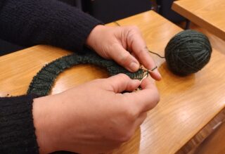 Käsissä neulontapuikot, pöydällä vieressä vihreää lankaa.