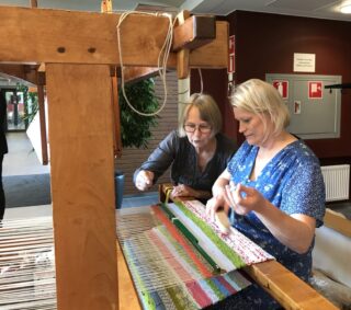 Kaksi naista kangaspuiden ääressä. Taaempi nainen opastaa toista. Värikästä mattoa näkyy jo kangaspuissa.