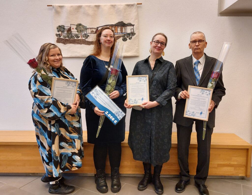 Neljä henkilöä vierekkäin seisomassa. Kolmella on kädessään kehystetty kunniakirja ja yhdellä palkintosummaa esittävä laminoitu julkaisu. Kolmella henkilöllä on myös punainen ruusu.