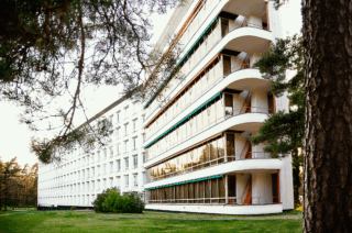 Suuri valkoinen rakennus kuvattuna metsän suunnasta.