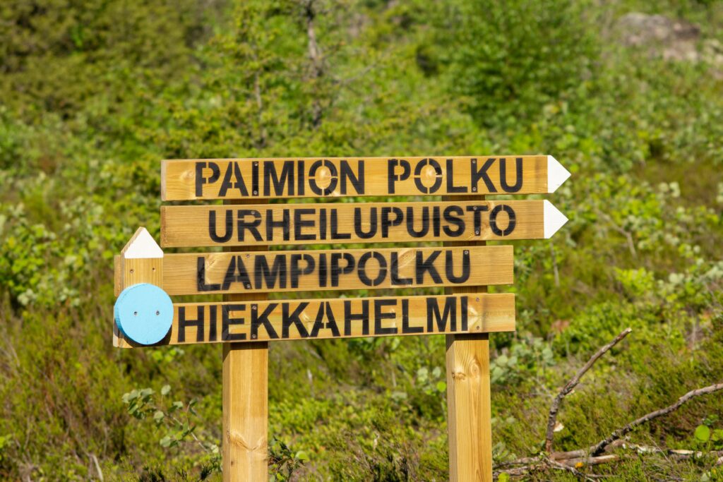 Vihreän luonnon keskellä oleva puinen kylttikokonaisuus, jonka kyltit osoittavat kahteen eri suuntaan.