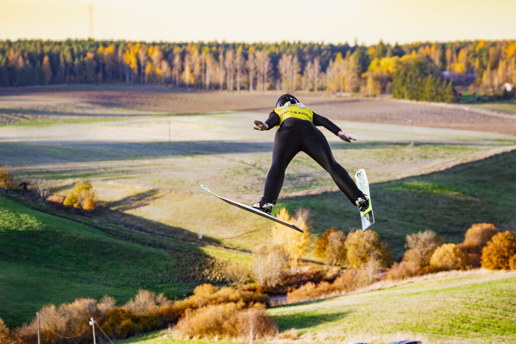 Takaapäin kuvattu henkilö, liitelee yläilmoissa irtauduttaaen mäkihyppytornin hyppyristä. Taustalla syksyinen aurinkoinen maisema.