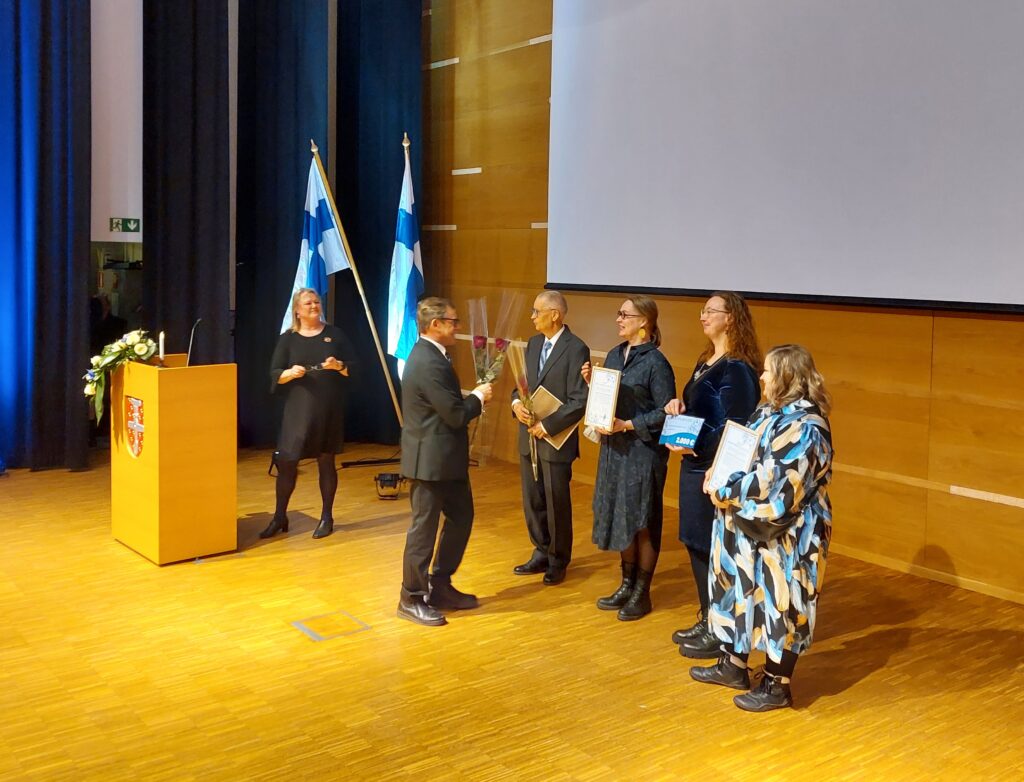Juhlasalissa kuusi henkilöä, joista neljä rivissä. Yksi henkilö jakaa ruusuja ja yksi seisoo puhujanpöntön takana. Taustalla näkyy kaksi Suomen lippua.