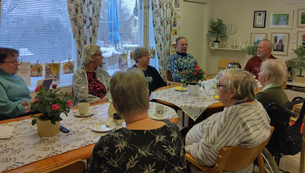 Pöytien ympärillä istuu eläkeikäisiä henkilöitä. Pöydällä muutamia ruukkuja ja kahvikuppeja.
