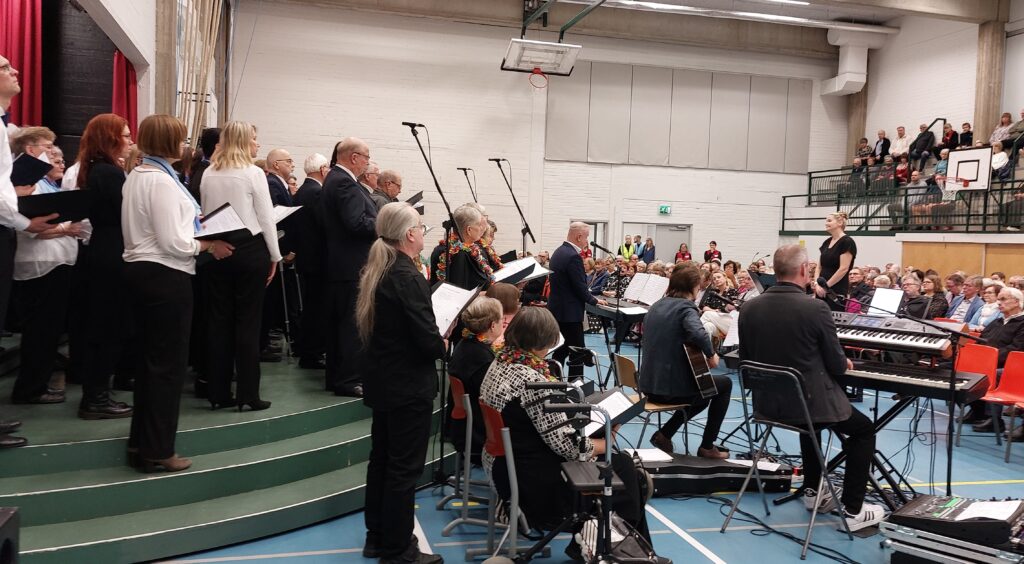 Liikuntasalissa esiintymässä iso kuoro, edessään solisti, muusikkoja ja kuoronjohtaja. Takana näkyy myös runsaasti yleisöä.