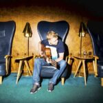 Mies istuu nojatuolissa ja soittaa kitaraa.