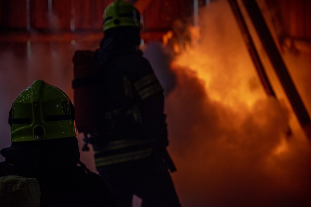 Kaksi pelastuslaitoksen työntekijää työvarusteissa etualalla. Taustalla voimakkaasti palava rakennus.