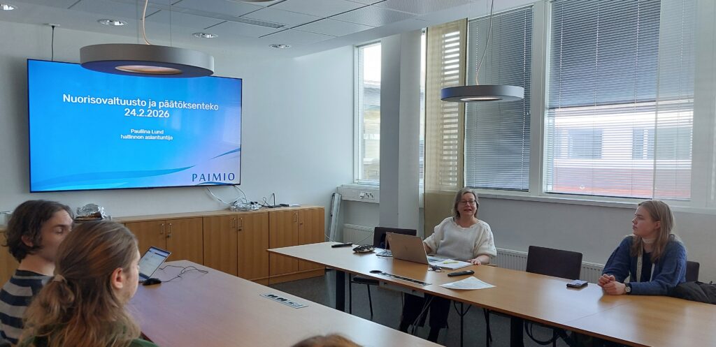 Henkilöitä istumassa kokoushuoneessa. Seinän näytöllä näkyy teksti Nuorisovaltuusto ja päätöksenteko.