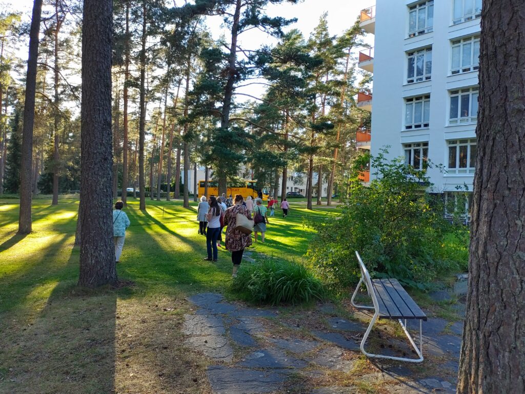 Kesäinen kuva metsikköalueelta. Oikeassa reunassa valkoinen rakennus ja taustalla keltainen linja-auto.