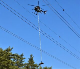 Takana helikopteri, etualalla sähkölinjoja, oikealla alhaalla puun latvoja.