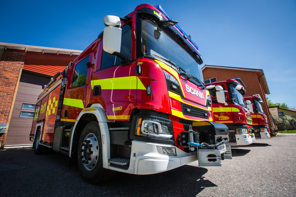 Kolme punaista paloautoa ulkona piha-alueella punertavan paloasemarakennuksen edustalla.