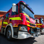 Kolme punaista paloautoa ulkona piha-alueella punertavan paloasemarakennuksen edustalla.