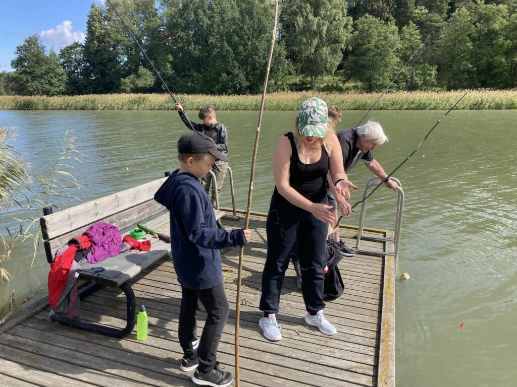 Eri-ikäisiä onkijoita laiturilla kesäisessä, aurinkoisessa säässä.