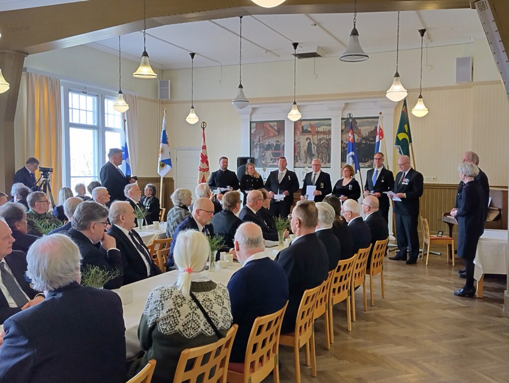 Juhlasali täynnä pöydissä istuvia vieraita. Salin etuosassa jääkärimitaleilla palkitut henkilöt seisovat rivissä.