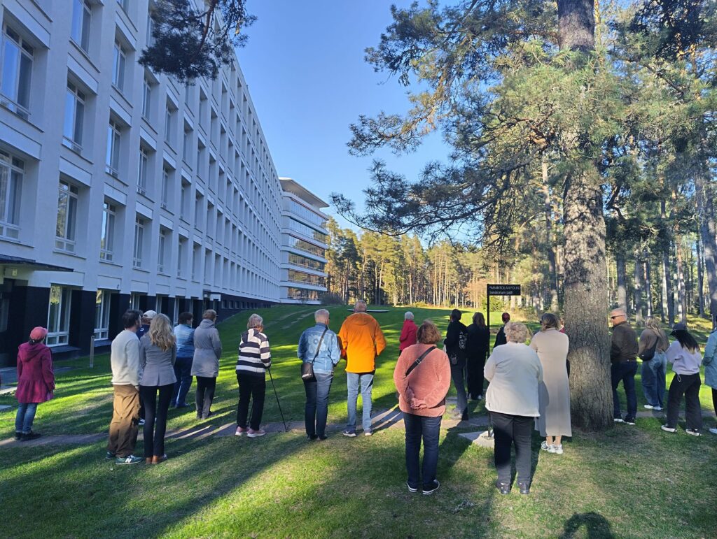 Ihmisiä seisoo vihreällä nurmikolla valkoisen kerrostalomaisen rakennuksen ulkoseinän tuntumassa.