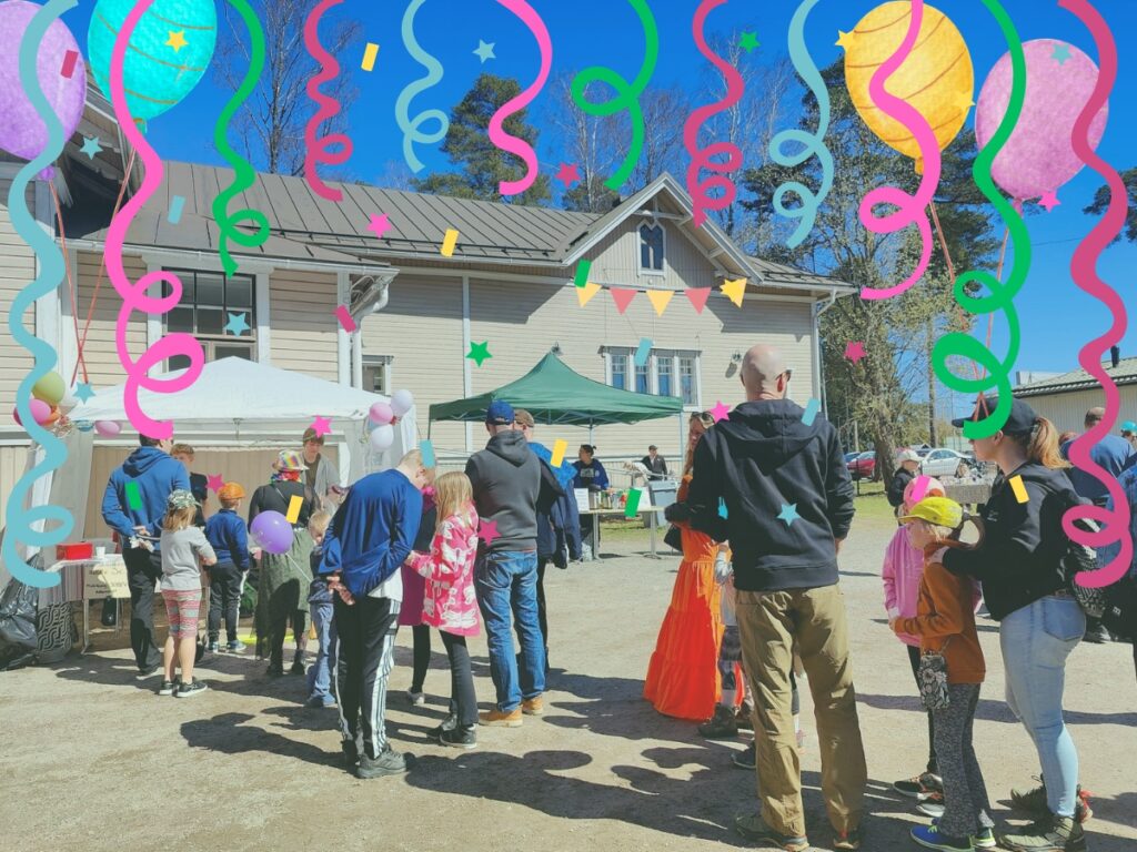 Eri-ikäisiä ihmisiä kauniissa kevätsäässä hiekkaisella pihalla, vaaleahkon rakennuksen edessä. Kuvassa näkyy myös ilmapalloja ja katoksellisia telttoja.