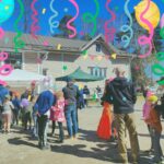 Eri-ikäisiä ihmisiä kauniissa kevätsäässä hiekkaisella pihalla, vaaleahkon rakennuksen edessä. Kuvassa näkyy myös ilmapalloja ja katoksellisia telttoja.