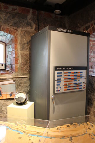 Taustalla suuri vanha tietokone, keskellä mittari jalustalla ja etualalla osa pienoismallia.