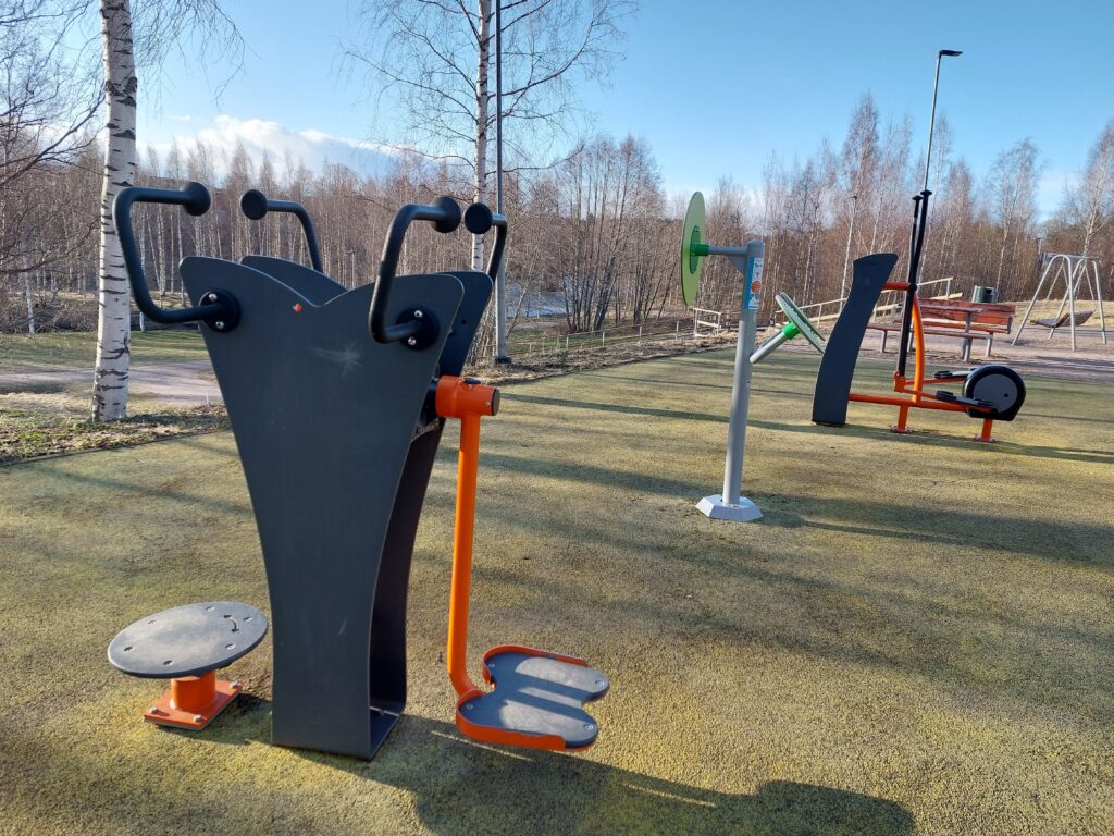 Etualalla ulkokuntosalilaitteita, taustalla keväisiä lehtipuita ja joki.