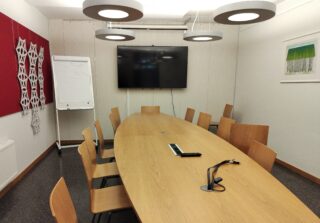 Kokoushuone, jossa etualalla näkyy iso pöytä ja tuoleja, taustalla fläppitaulu ja tv-näyttö. Yhdellä seinällä myös maalaus.