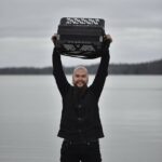 Tummaan asuun pukeutunut mies seisoo rannalla ja pitää päänsä päällä haitaria.