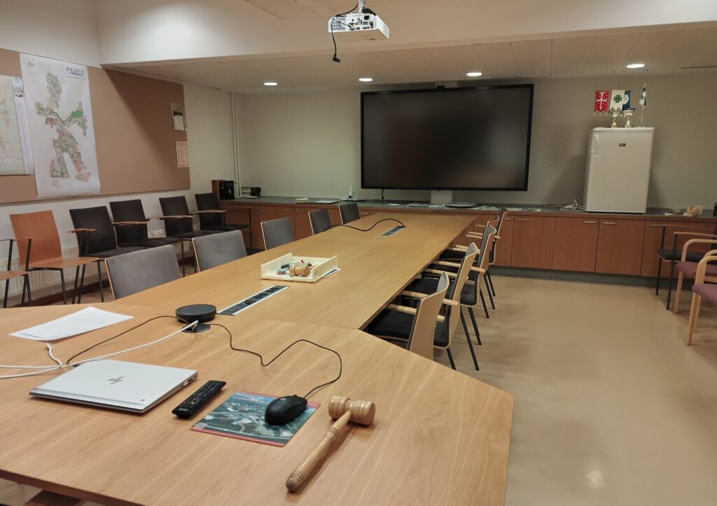 Etualalla iso kokouspöytä, jonka päällä kannettava tietokone, puheenjohtajan nuija, kaukosäädin ja kyniä. Takaseinällä on iso tv-näyttö ja katossa videotykki.