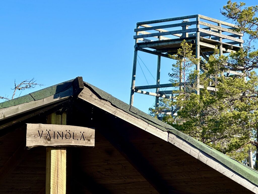 Taustalla puinen näkötorni ja etualalla laavun katos, johon kiinnitetty puinen kyltti, jossa lukee Väinölä.