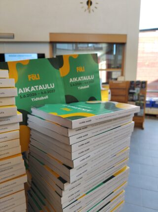 Pehmeäkantisia paljon päällekkäin aulatilassa olevalla pöydällä. Taustalla näkyy esiteteline ja seinäkello.