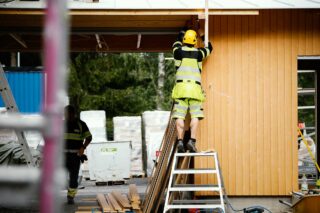 Kypäräpäinen rakennusmies on ulkona asentamassa telineen päällä rakennukseen seinään jotakin.