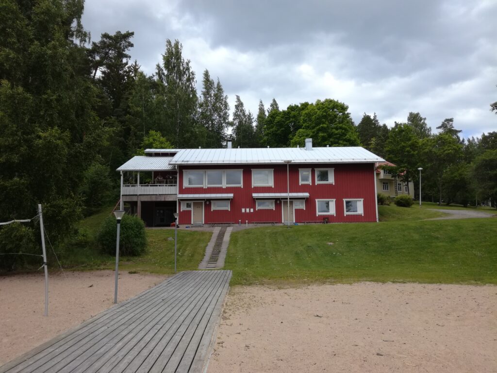 Rinteessä oleva, punertava kaksikerroksinen rakennus, joka on kuvattuna puolipäiväisenä kesäpäivänä. Etualalla hiekkaa ja laatoitettu polku.
