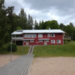 Rinteessä oleva, punertava kaksikerroksinen rakennus, joka on kuvattuna puolipäiväisenä kesäpäivänä. Etualalla hiekkaa ja laatoitettu polku.
