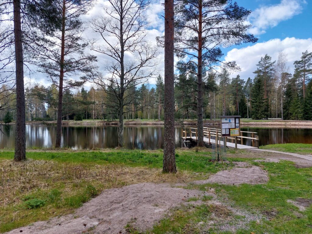 Etualalla ranta-alueella muutama puu ja nurmikkoa, niiden takana uimalampi ja sen takaa enemmän puita.