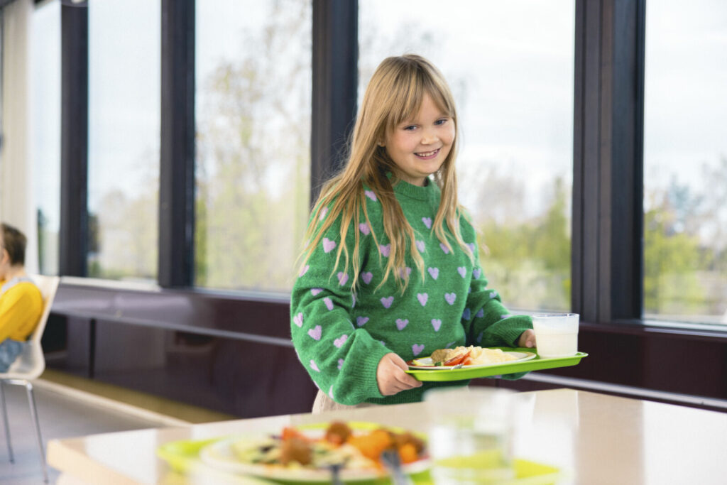 Ruokalan linjastolla vihreäpuseroinen, hymyilevä tyttö kuljettaa ruoka-annosta tarjottimella.