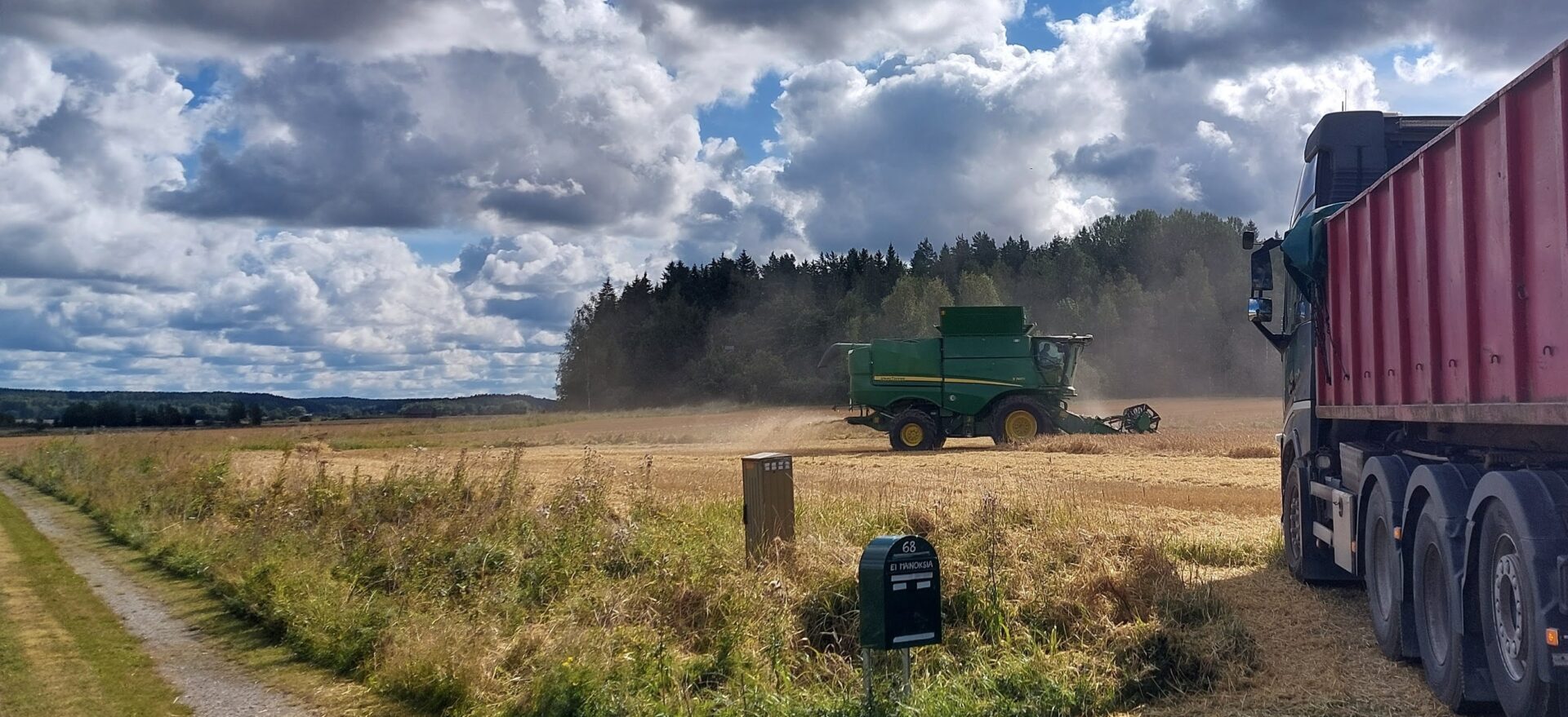 Loppukesän kuvassa puimuri puimassa pellolla puolipilvisessä säässä. Etualalla näkyy kuormalava-auto ja postilaatikko.