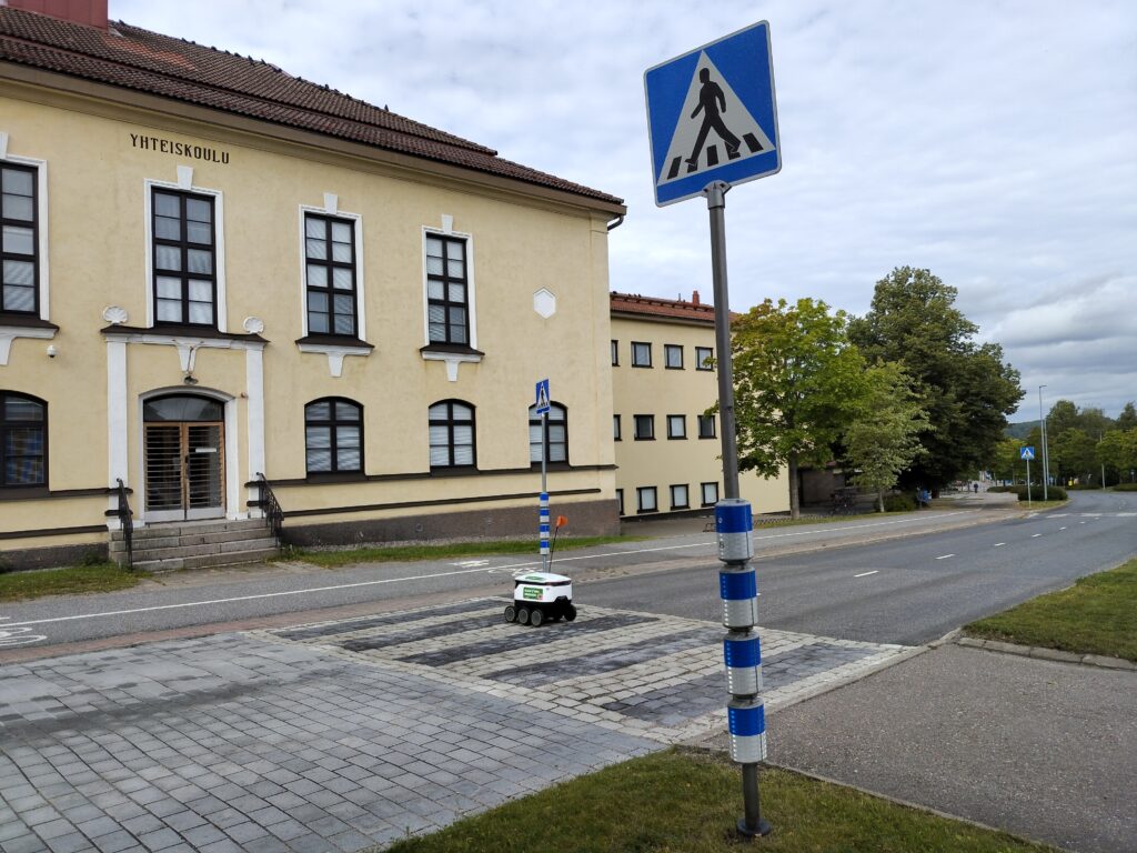 Katu, jonka yli menee suojatie. Suojatie-liikennemerkkeihin lisätty välkkylaitteisto. Taustalla kellartävä kivirkannus, jonka seinässä lukee Yhteiskoulu.