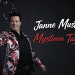 Tumman taustan edessä monivärisessä puvuntakissa oleva tummatukkainen mies.